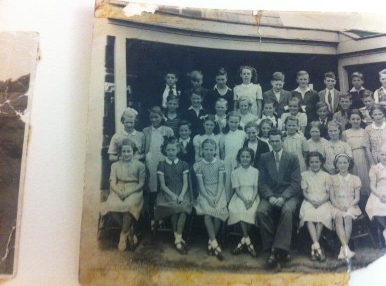 A photograph of a school photograph, partly torn at the corners: a class of about forty boys and girls in rows in front of a verandah, with a male teacher in a suit seated in the middle of the front row. The edge of another print shows at the left.