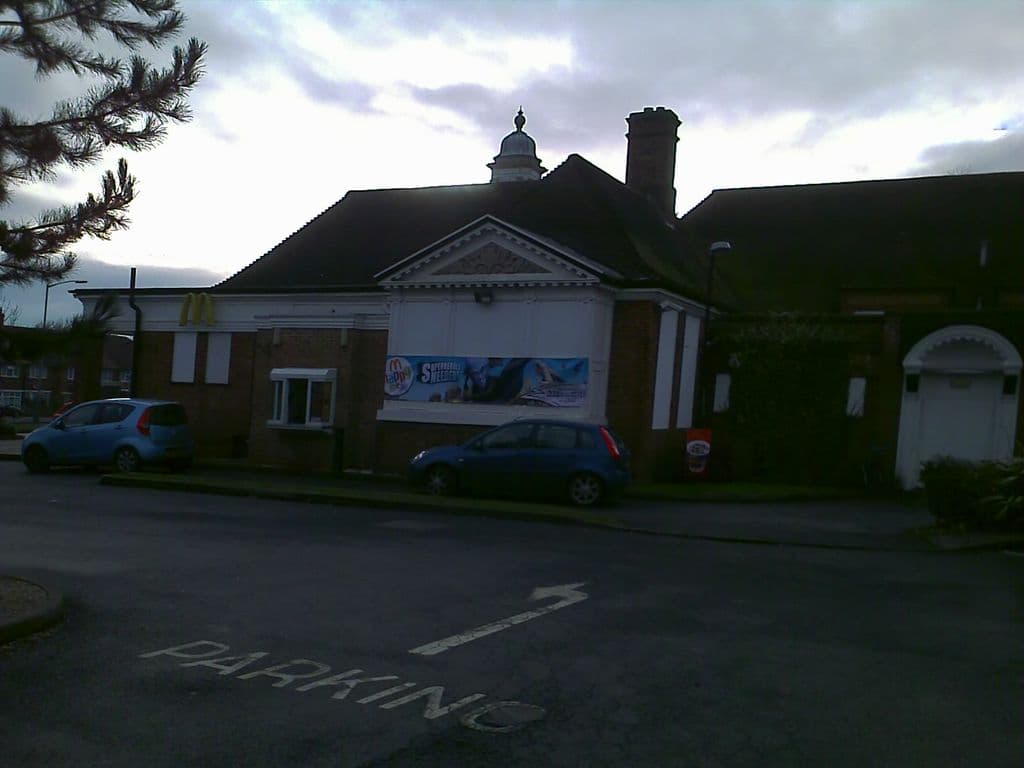 2010: The former College Arms pub, a long brick building with a tiled roof, a small cupola and a white pedimented entrance, now carrying McDonald’s golden arches and a promotional banner; cars are parked in front and “PARKING” is painted on the road.