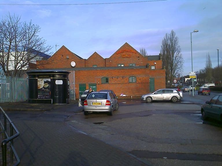 A long red-brick pub building with three gables, the middle one lettered “Kingstanding” and “M&B”, behind a small car park with a public toilet kiosk on the left and cars on the forecourt.