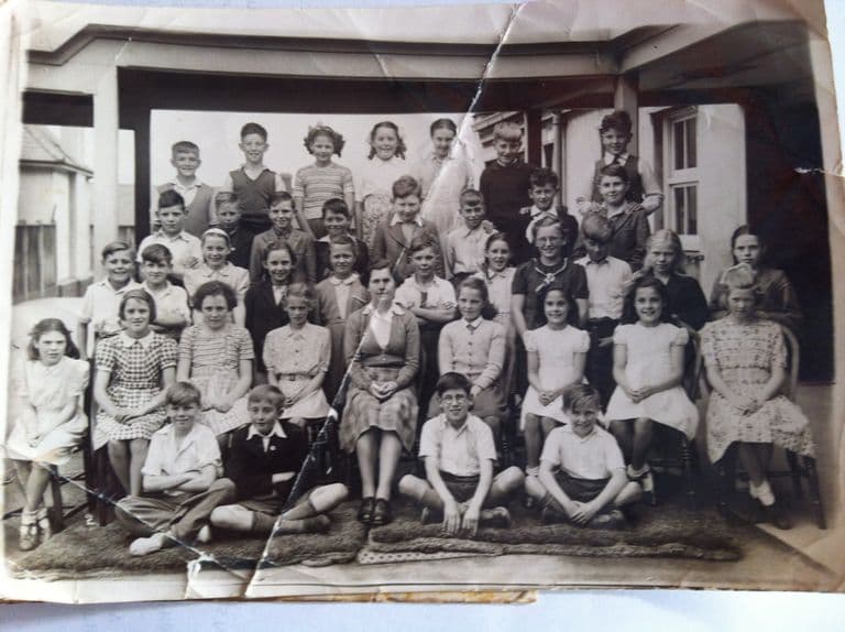 A creased and cracked class photograph: about thirty-five children in four rows under a verandah, the boys at the front sitting cross-legged on mats and a woman teacher seated in the centre.