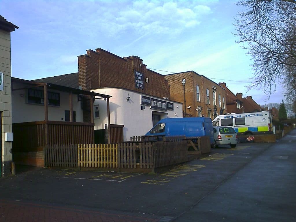 2010: A white-painted single-storey function-room building with a sign reading “Function Room Available”, a covered wooden smoking area and picket fence in front, a taller brick building behind and a police van parked by the neighbouring police station.