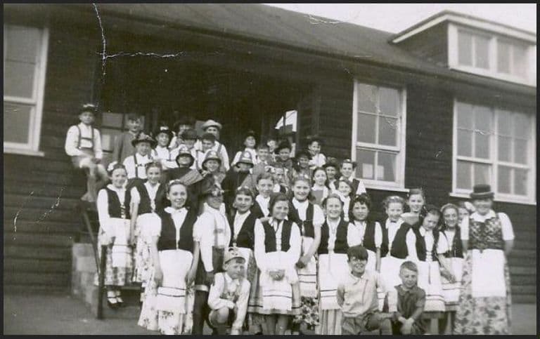 About forty children in Alpine-style costume – girls in dirndls, white blouses and aprons, boys in braces and hats – crowd on the steps of a wooden classroom block.