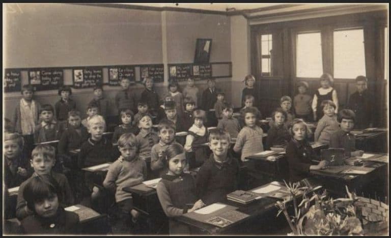 A sepia classroom photograph: about forty young children at wooden double desks turn towards the camera, with reading cards along the wall, tall windows on the right and a vase of flowers on the teacher’s desk in the foreground.