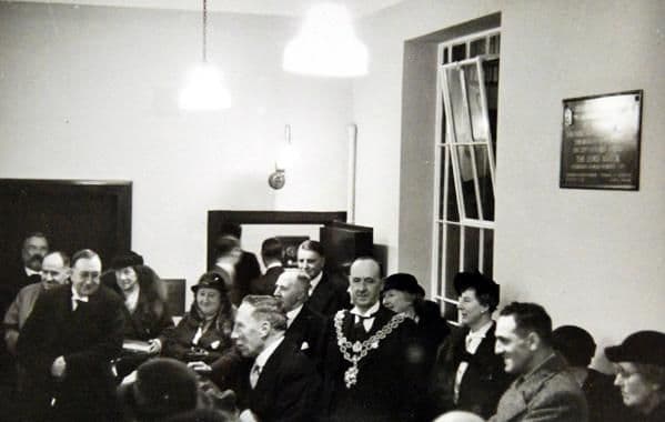 Inside the new bank, men in overcoats and women in hats stand round the Lord Mayor, who wears his chain of office; a commemorative plaque is on the wall and pendant lamps hang from the ceiling.