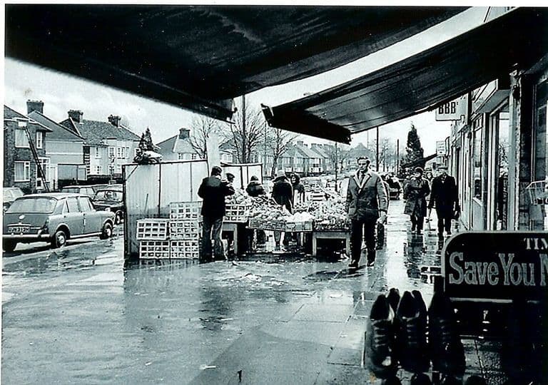 Looking along a rain-soaked pavement from under shop awnings: a greengrocer’s stall piled with produce and crates, shoppers in coats, a parked 1960s saloon car, semi-detached houses across the road and a shoe-shop sign reading “Save You M…” in the foreground.