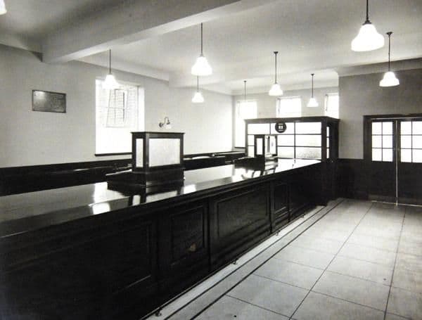 The empty banking hall of the new branch: a long polished wooden counter with a glazed screen, a tiled floor, pendant lamps, a plaque on the wall and a partly glazed office at the far end.
