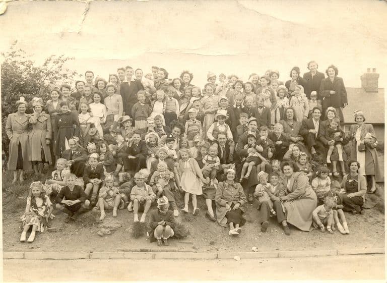 A large crowd of neighbours – women in coats and hats, men, and children in paper crowns and fancy dress – posed on a grassy bank above a kerb, with a house chimney at the right.