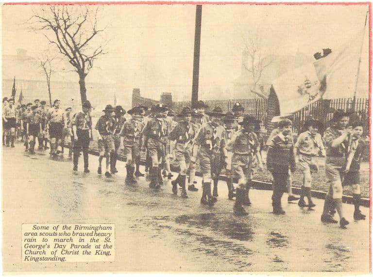 A newspaper photograph of Scouts in uniform and broad-brimmed hats marching along a wet road past railings, with a flag carried at the front. The printed caption reads “Some of the Birmingham area scouts who braved heavy rain to march in the St. George’s Day Parade at the Church of Christ the King, Kingstanding.”