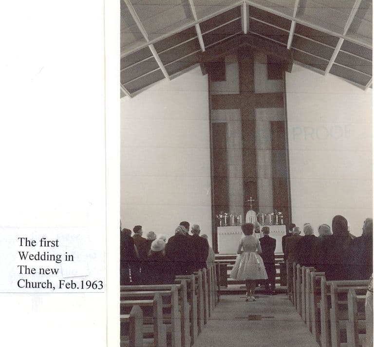 The interior of the new church, looking down the aisle between rows of pews to a bride and groom at the altar under a tall timber cross and a steep boarded roof; a label reads “The first Wedding in The new Church, Feb. 1963”.
