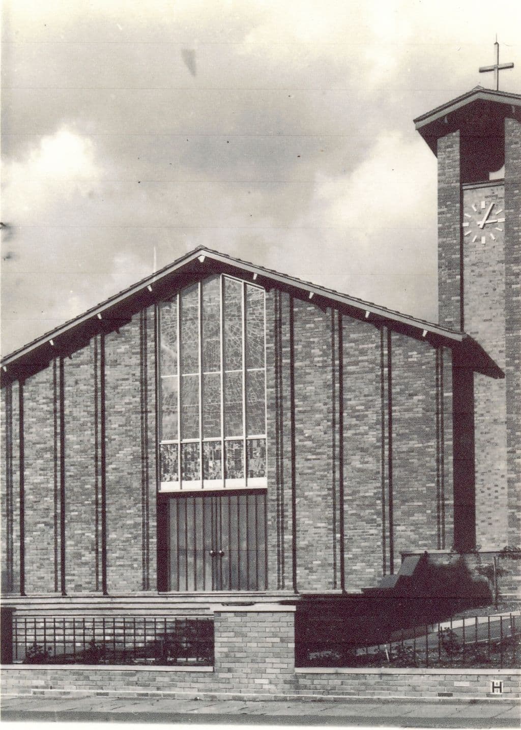 1962: A black-and-white view of the new church’s brick west front: a tall gable with a large stained-glass window above double doors, a clock tower with a cross on the right, and a low wall with railings in front.