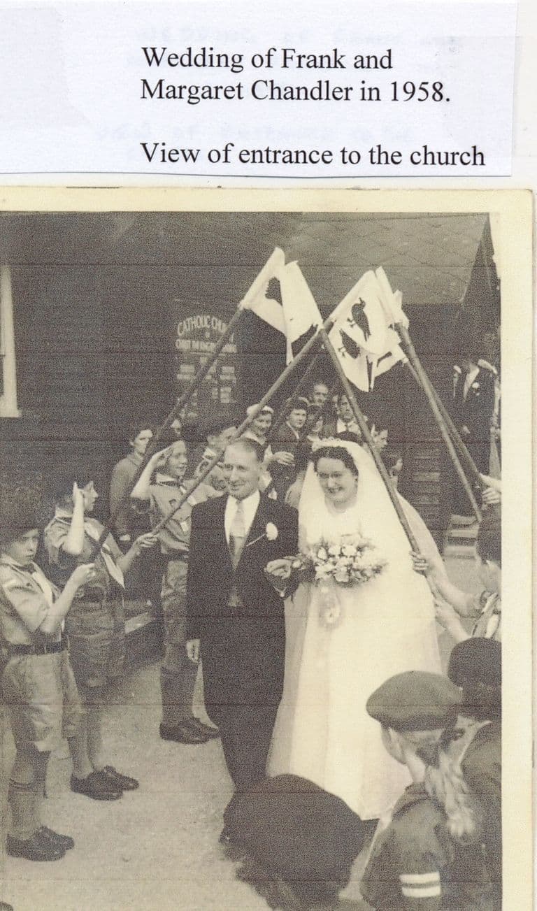 A bride and groom leave a wooden church under an arch of flags on staves held by scouts and cubs; a noticeboard behind reads “Catholic Church of Christ the King”. A label above says “Wedding of Frank and Margaret Chandler in 1958. View of entrance to the church”.