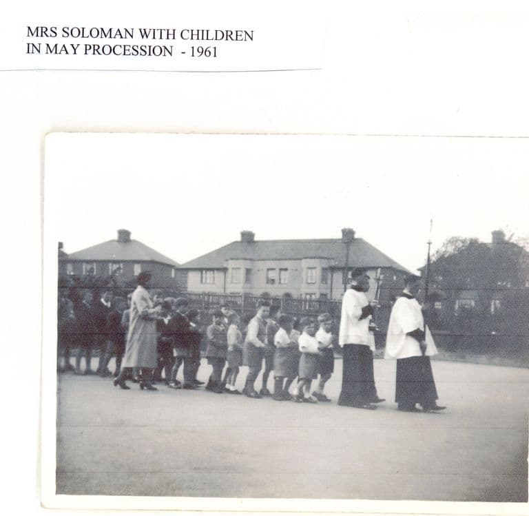 A line of small children led by two altar servers in cassocks and surplices, one carrying a cross, walks across a playground with a woman teacher, with 1930s semi-detached houses behind; the label reads “Mrs Soloman with children in May procession – 1961”.