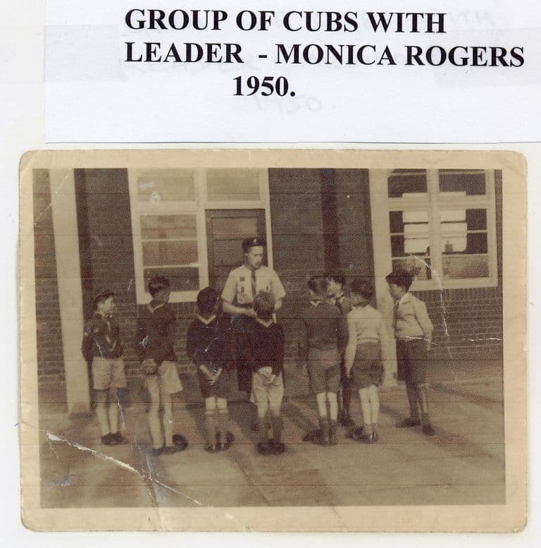 A worn snapshot of about ten small boys in cub uniform standing with their backs to the camera round their leader, in front of a brick building with large windows; a typed label reads “Group of cubs with leader – Monica Rogers 1950.”