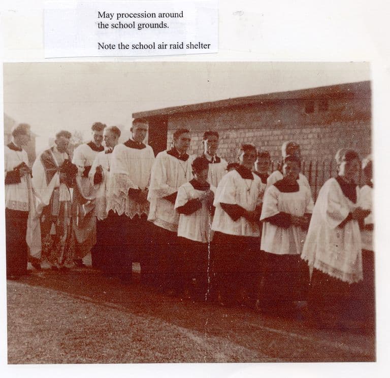 A sepia photograph of a line of altar servers and men in white lace surplices standing on a gravel path in front of a low brick building; a label above reads “May procession around the school grounds. Note the school air raid shelter”.
