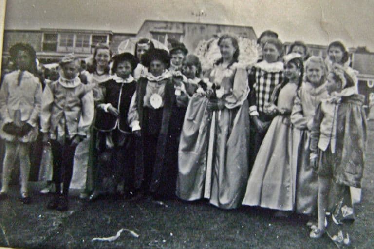Children in Tudor-style pageant costume – ruffs, long satin gowns, a crowned “queen” with a large fan-like collar – stand on grass in front of the school’s flat-roofed buildings.