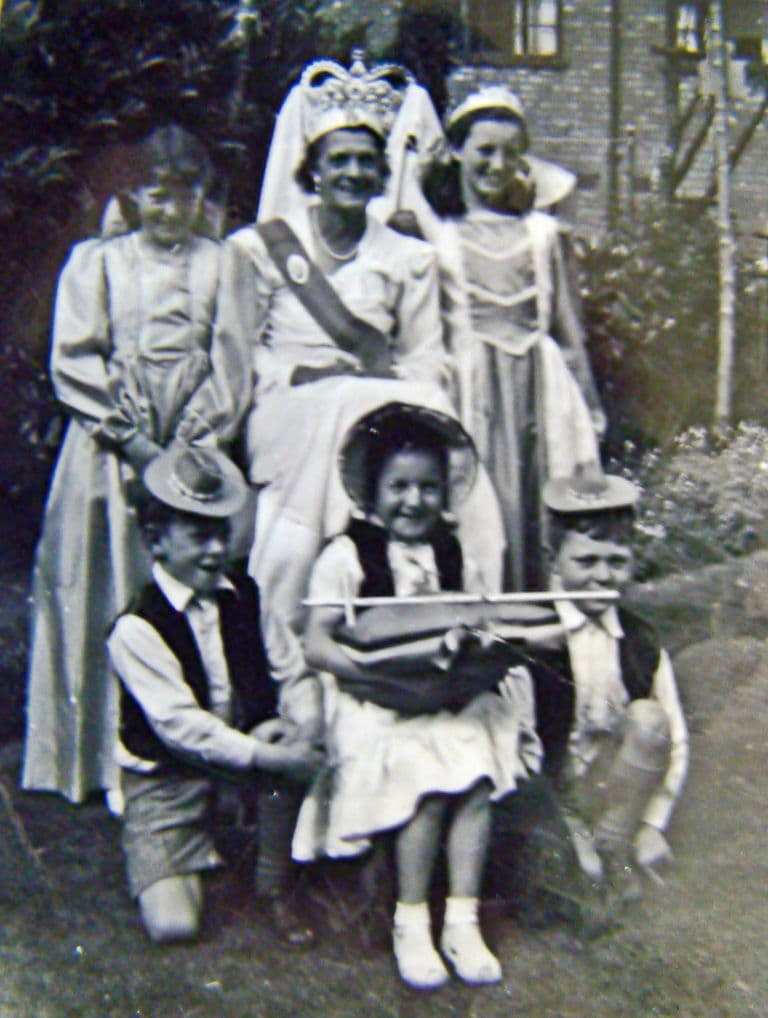 Six children in fancy dress in a back garden: a girl crowned as the Queen with a sash stands between two girls in long gowns, and in front three younger children in cowboy hats and waistcoats, the girl holding a toy crossbow.