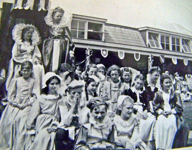 Girls in Elizabethan costume with ruffs and headdresses crowd together on and around a decorated float or stage, two standing high at the back, in front of the school’s long windowed buildings hung with heraldic shields.