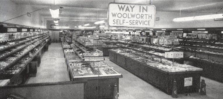 The inside of a Woolworth store: long island counters and wall shelves stacked with goods, strip lights overhead and a hanging sign reading “Way in – Woolworth self-service”.