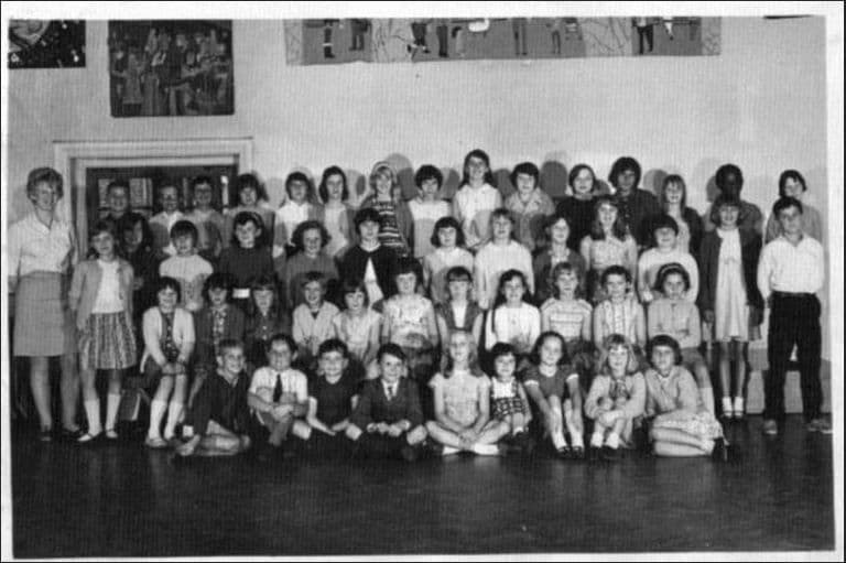 About fifty children in rows, some seated cross-legged on the floor, in a school hall with children’s paintings on the wall; a woman teacher stands at the left and a boy at the right.