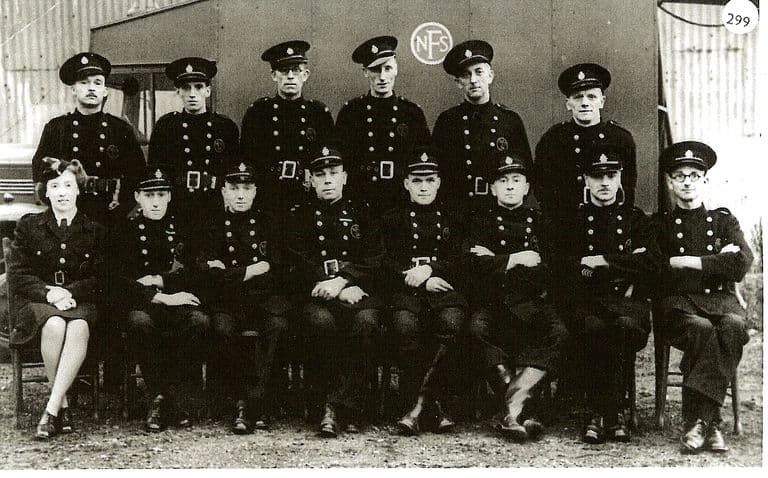 Fourteen firemen in double-breasted tunics, belts and peaked caps and one woman officer in uniform, posed in two rows in front of a hut marked “NFS”; a lorry stands at the left. A circled “299” is printed in the top corner.