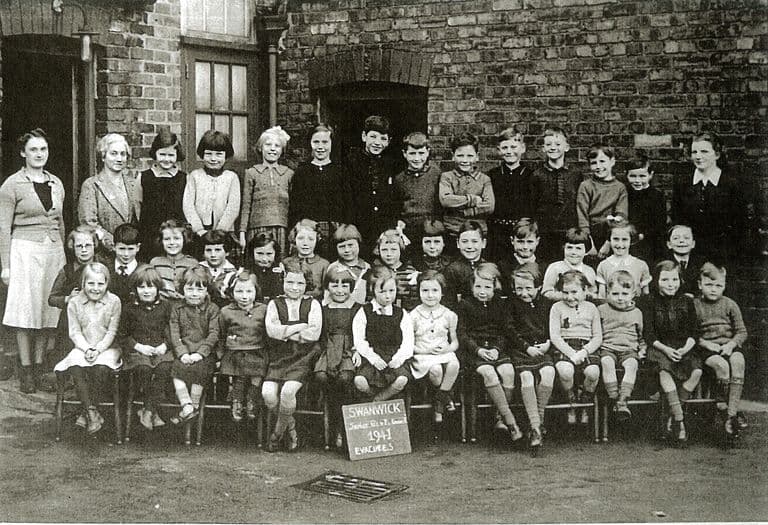A school group of about forty children in three rows, with three women teachers, in front of a brick school building. A slate on the ground reads “Swanwick … 1941 Evacuees”.