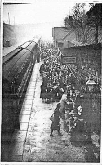 A small, grainy copy of a crowded railway platform: a long line of children and adults with luggage beside a train, a station building and a lamp on the right.