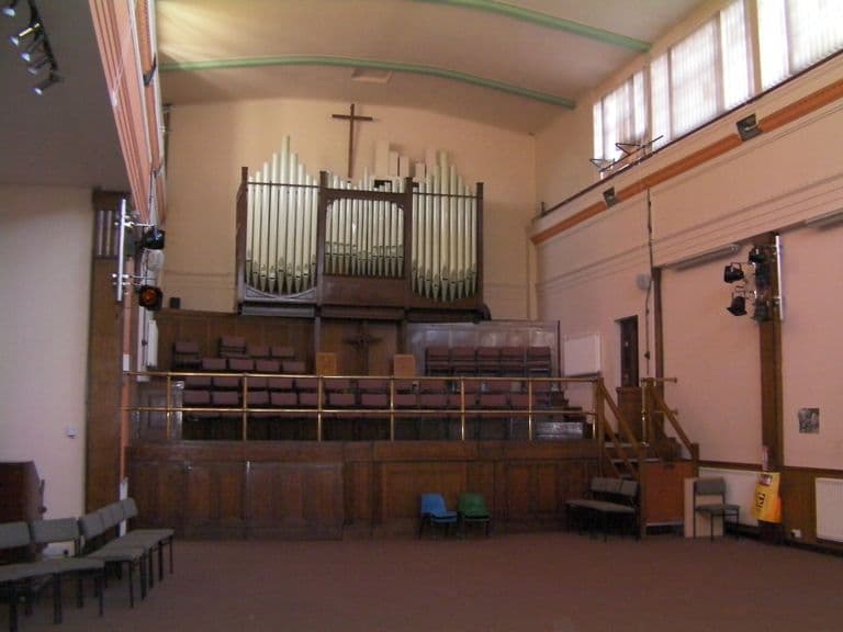 The inside of a tall church hall with a large pipe organ and a wooden cross above tiered choir seating behind a brass rail, stage lights on the walls and rows of chairs on the carpet.