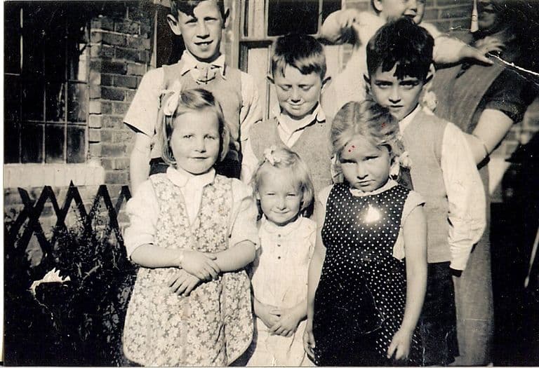 Black-and-white snapshot of three young girls in summer frocks standing in front of three older boys in knitted tank tops, outside a brick house with a trellis fence; an adult and a baby are partly visible behind.