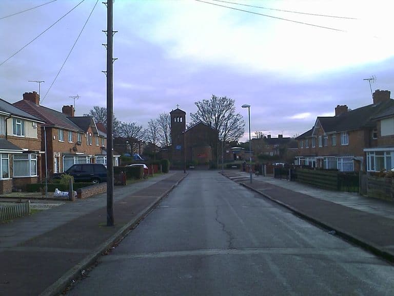Looking down a quiet street of 1930s brick semi-detached houses towards St Luke’s Church, a brick building with a square bell tower topped by a cross, among bare trees.