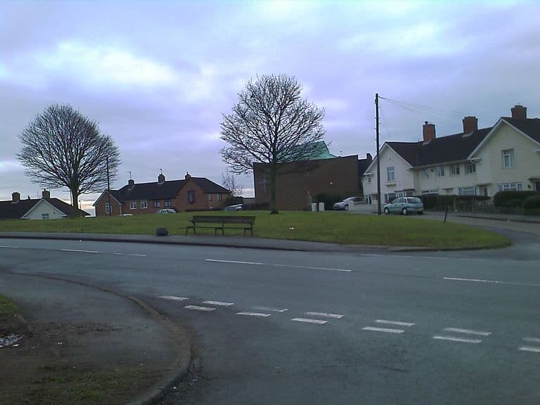 A grassy green with a bench and bare trees at a road junction; the brick St Mark’s church with its green copper roof stands behind, between rows of houses.