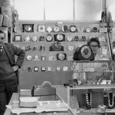 Black-and-white photo inside a jeweller’s shop: shelves of mantel clocks and alarm clocks fill the back wall, a glass counter holds necklaces and watches, a man in a jacket stands at the left and a woman in glasses behind the counter.