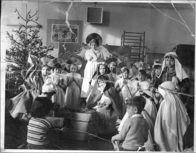 Black-and-white photograph of young children in a nativity play in a school hall: angels with haloes and tinsel, shepherds in head-dresses, Mary with a doll by a straw-filled crib and a Christmas tree on the left; the print is cracked.