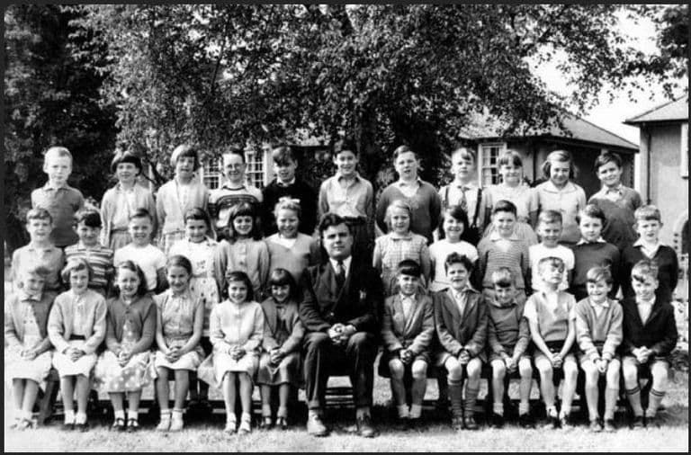 Black-and-white class photograph of about thirty children in three rows on grass under a large tree, a male teacher in a dark suit seated in the centre, school buildings behind.