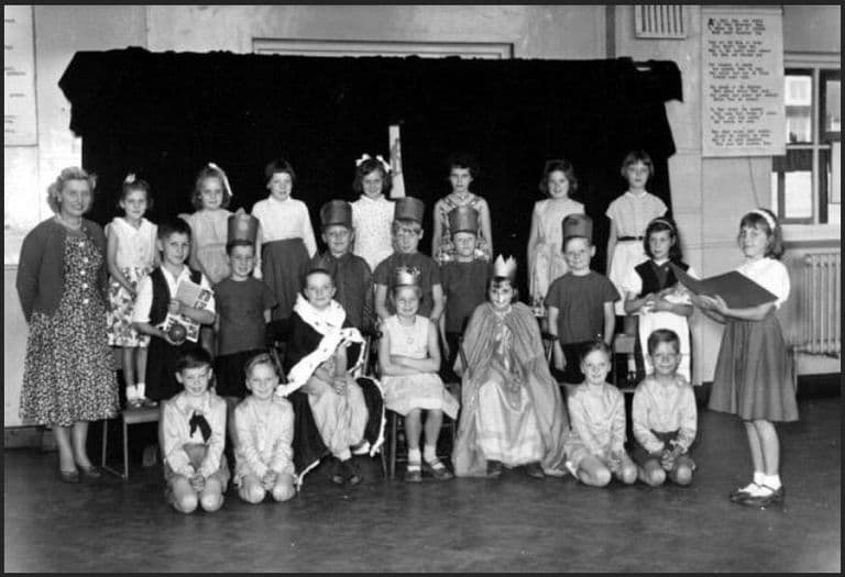 Black-and-white photograph of a class in costume in a school hall: a crowned king and queen seated in the middle, boys in paper hats, girls in party frocks, a narrator holding a book at the right and a woman teacher at the left.
