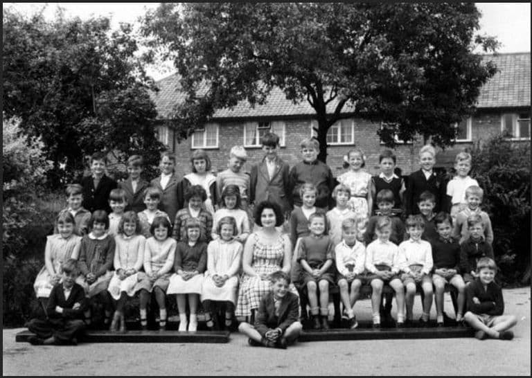 Black-and-white class photograph of about thirty-five children on benches in a yard, a dark-haired woman teacher in a checked dress seated in the centre, a tree and a brick house with a tiled roof behind.