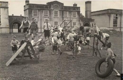 Young boys play on wooden PE apparatus on the school field – sloping ladders, a climbing frame and a tyre – in front of a two-storey school building with a tall chimney.