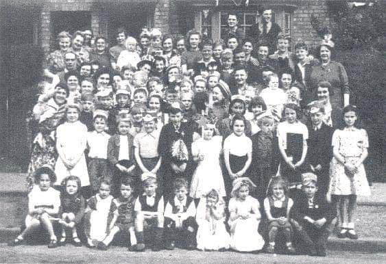 Black-and-white group photograph of dozens of children and adults posed in rows in front of a house, several children in fancy dress and paper hats.