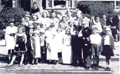 Small black-and-white group photograph of children in fancy dress and party hats standing on a pavement in front of houses and a hedge, with adults behind.