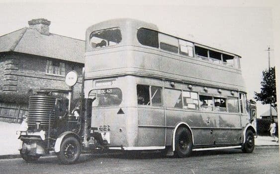 A grey double-decker bus, registration OG 421, towing a small two-wheeled trailer carrying a tall cylindrical gas-producer unit, parked on a road beside houses.