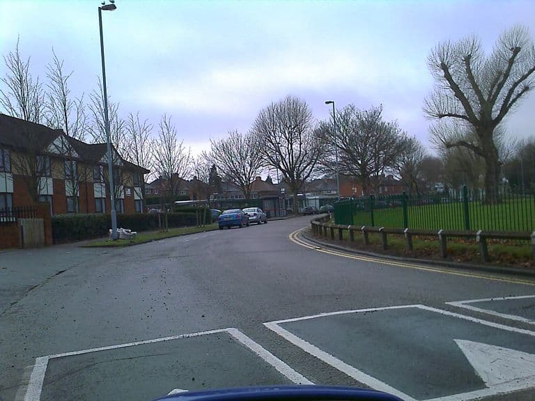 A curving road lined with bare trees; low school buildings sit behind green railings at the end of the road, with modern flats on the left and a playing field on the right.