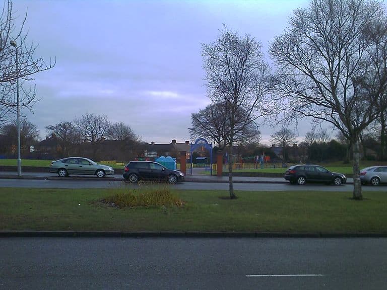 A grassed park with a children’s playground behind a blue arched gateway between brick pillars, seen across a road and a grass verge with young trees; houses line the far side.