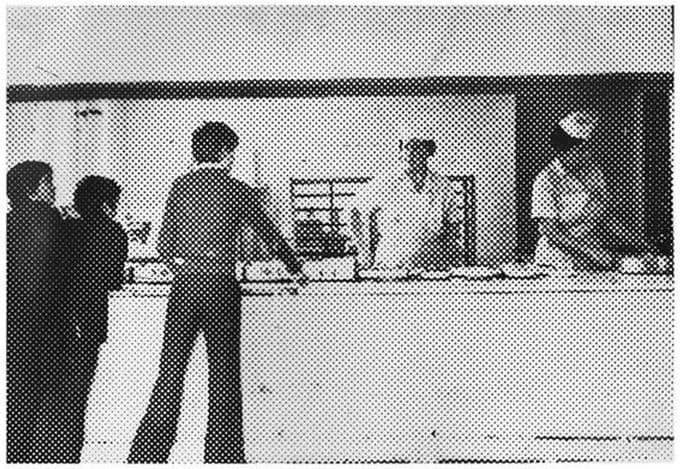 A coarse half-tone picture of pupils queuing at a long serving counter while dinner ladies in white overalls and caps serve behind it.