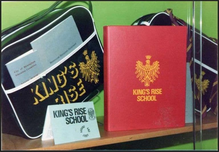 Colour photograph of a black holdall printed “King’s Rise” with a crowned eagle badge, a red folder with the same gold eagle, a City of Birmingham Education Committee note book and a small “King’s Rise School” card on a shelf.