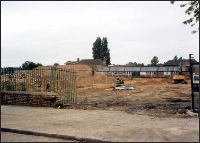 Colour photograph of a large cleared site of churned earth with a roller and a digger, behind a bent green fence and low wall; a long single-storey school block and houses stand at the back.