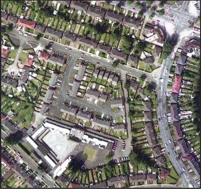 A vertical colour aerial view of streets of semi-detached houses with long back gardens and parked cars, with a flat-roofed school building and playground at the bottom left.