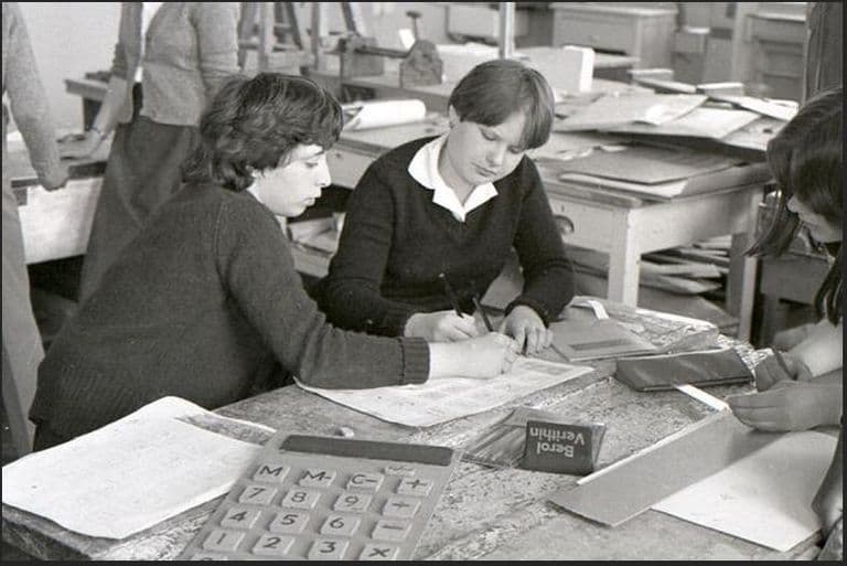 Black-and-white photo of two teenage girls in dark jumpers drawing on paper at a worn workbench, with a large calculator, a Berol Verithin pencil box and stacks of card around them.