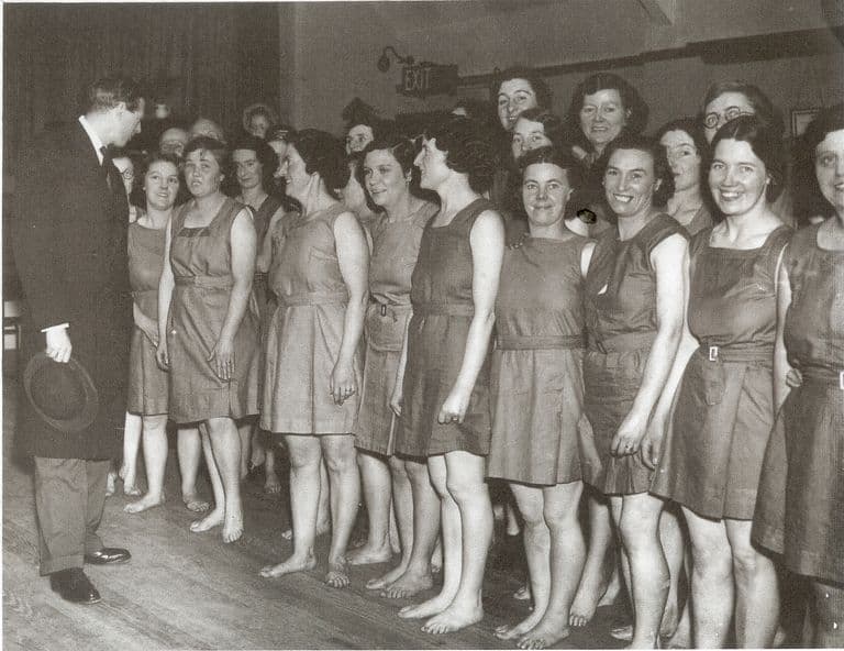 Black-and-white photograph of a line of smiling women in matching sleeveless belted tunics, barefoot on a wooden hall floor, being greeted by a man in a dark morning coat holding a hat; an EXIT sign is on the wall behind.