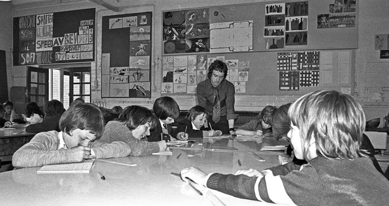 Black-and-white photo of boys and girls drawing at large white tables in an art room, a young male teacher in a patterned tie leaning over them; the walls are covered in pupils’ paintings, lettering designs and a football team picture.