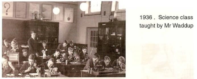 A scanned booklet page: boys at benches with scientific apparatus in a laboratory with glass-fronted cabinets and hanging lamps, a teacher standing among them. Printed beside it: “1936. Science class taught by Mr Waddup”.