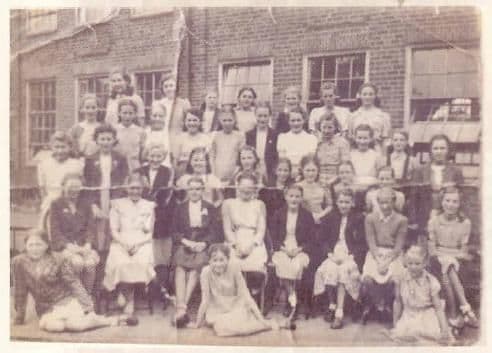 A creased class photograph of about thirty-five girls in four rows outside a brick school building, some seated on chairs and two sitting on the ground in front.